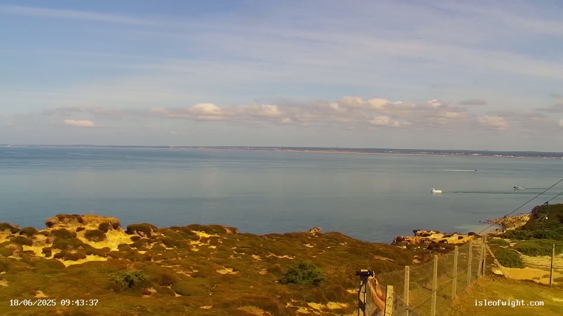 A calm sea stretches to a distant shoreline under a partly cloudy sky, viewed from a high vantage point overlooking scrubland and a low fence.