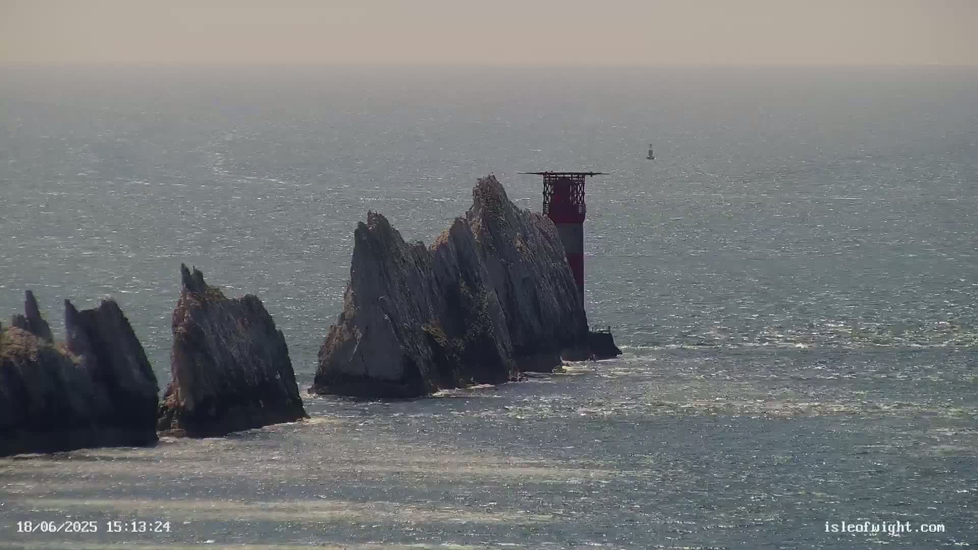 A red and white lighthouse stands atop a cluster of jagged, grey rock formations jutting out of a slightly choppy sea on a hazy day.