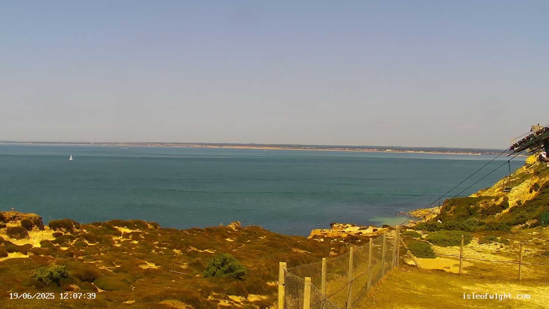 A sunny day reveals a tranquil ocean scene, viewed from a clifftop with sparse vegetation and a cable car system partially visible.