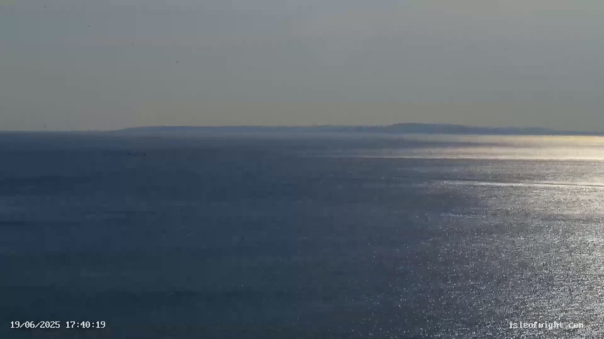 A calm, glistening ocean stretches to a hazy horizon line where land is barely visible under a bright, slightly overcast sky.