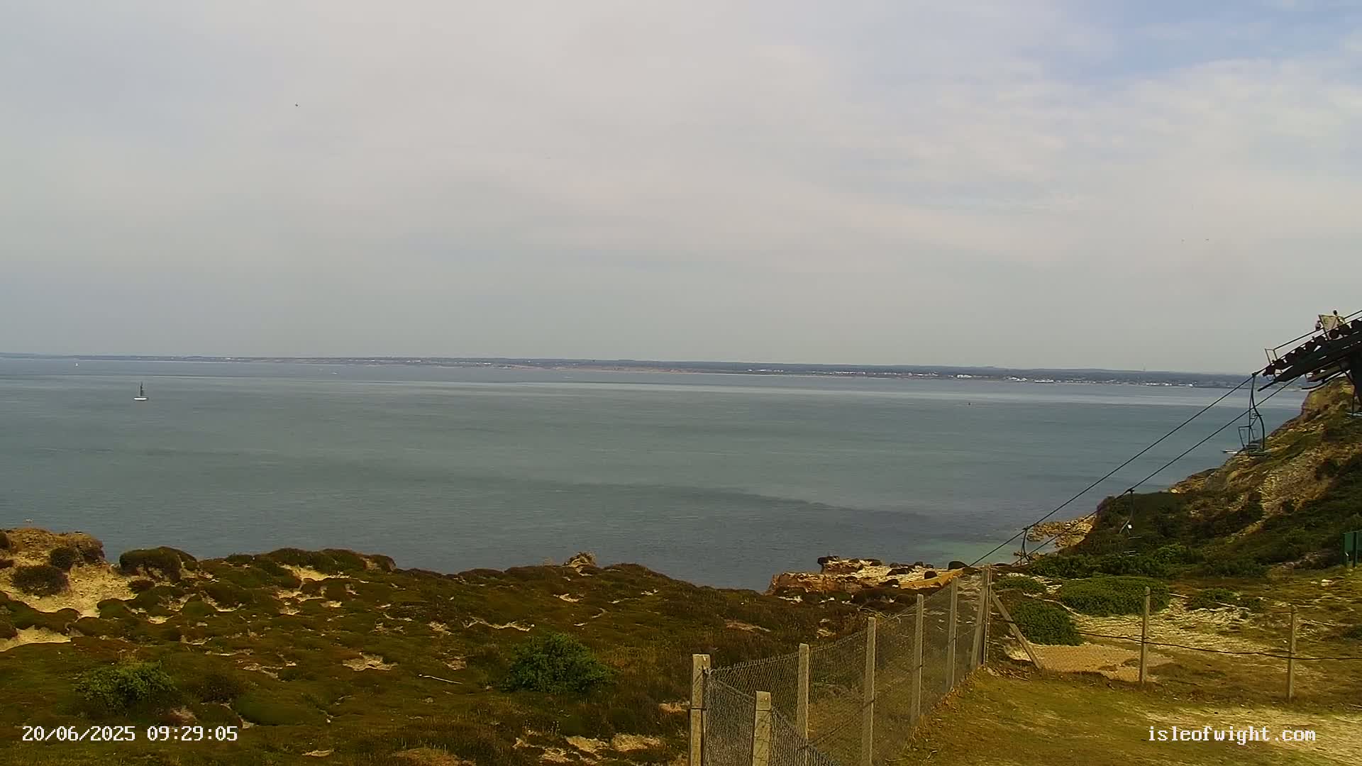 A calm, overcast day reveals a view of a wide expanse of ocean from a grassy clifftop, featuring a distant shoreline and a chairlift partially visible in the foreground.
