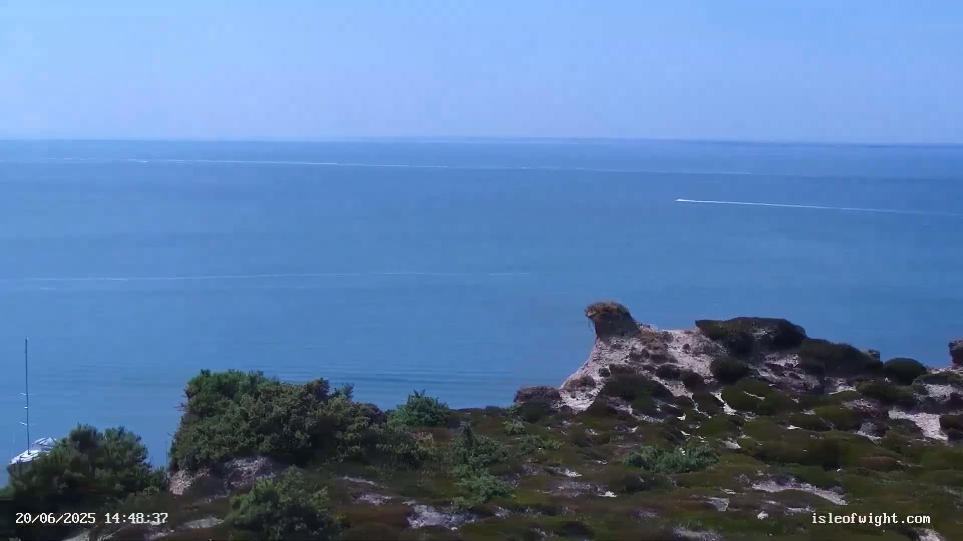 A calm, blue ocean stretches to the horizon from a high vantage point overlooking a rocky, shrub-covered coastline on a clear day.