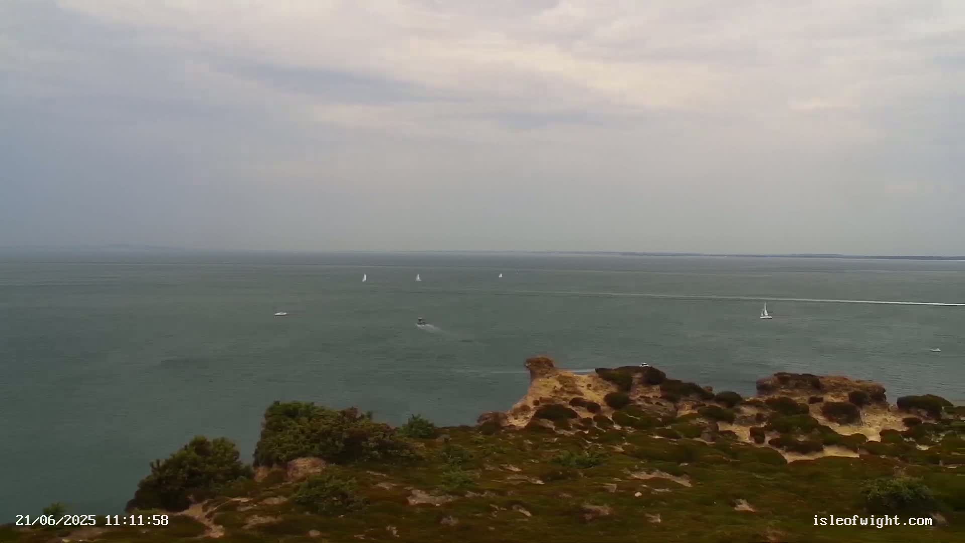 A coastal view shows a calm, gray sea with several sailboats and a motorboat, viewed from a clifftop covered in low, green vegetation under an overcast sky.