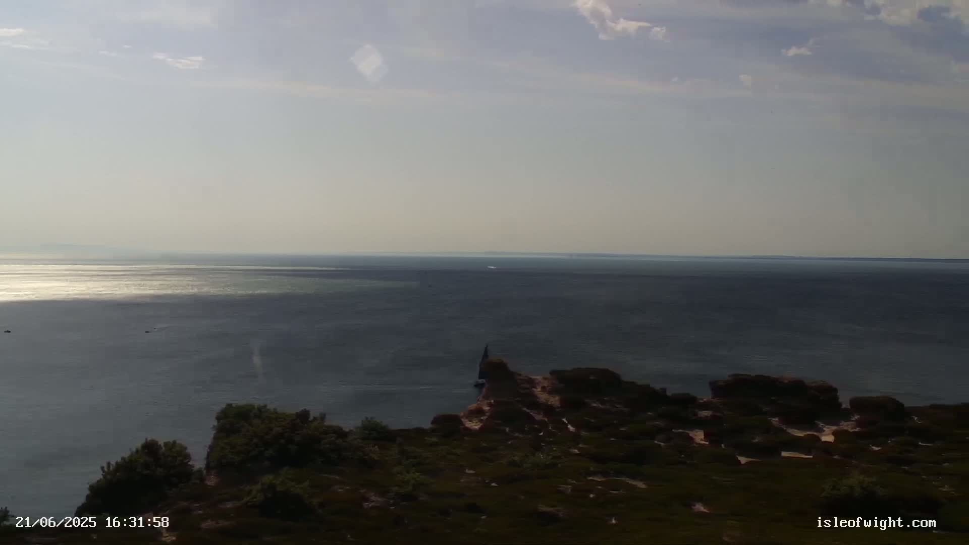 A high-angle view shows a calm sea under a mostly clear sky, with a small rocky outcrop and low-lying vegetation in the foreground.