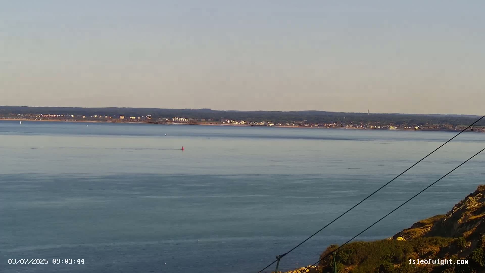 A calm, expansive body of water stretches to a distant shoreline under a clear sky, viewed from a high vantage point on a rocky outcrop.