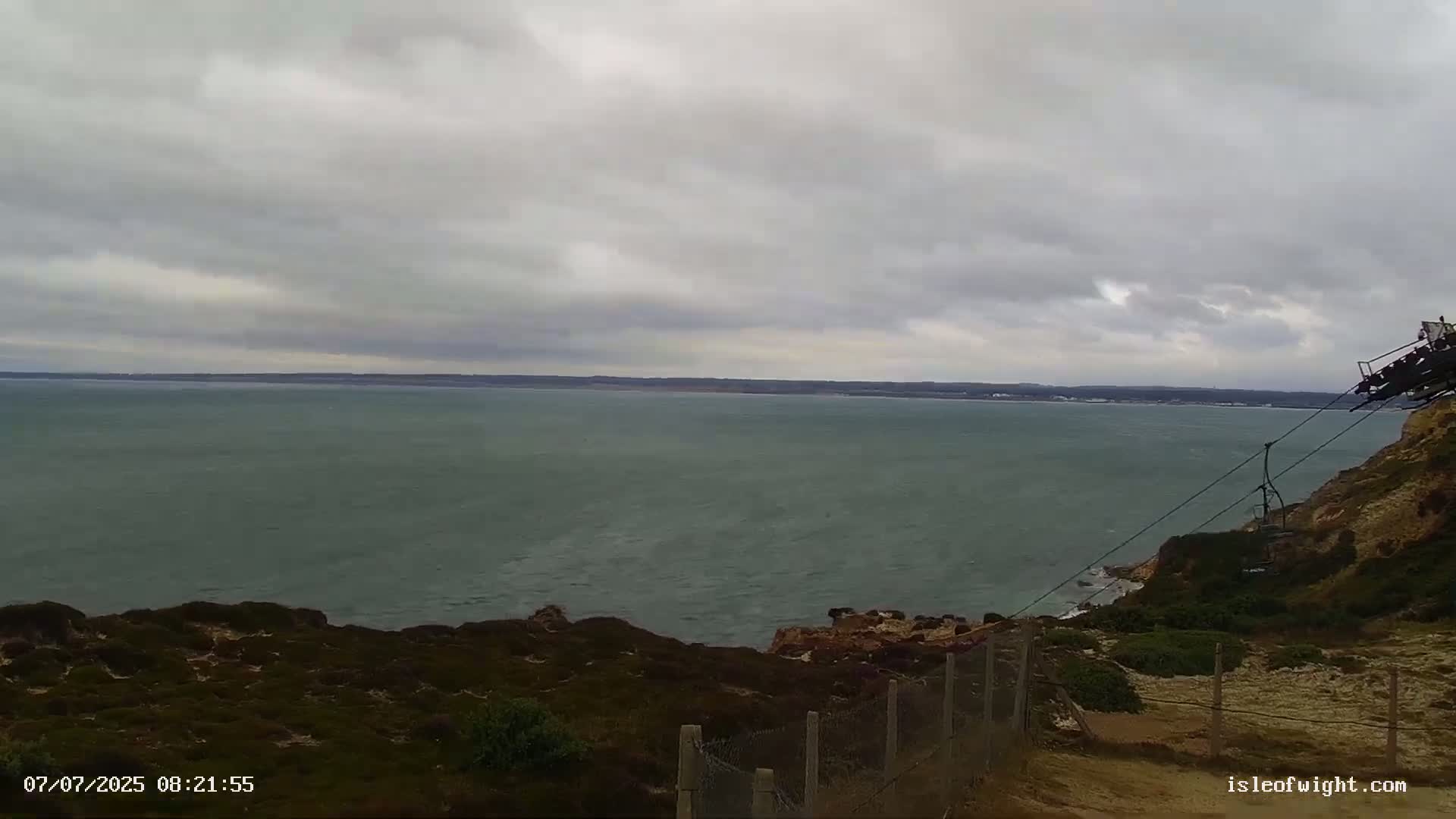 A mostly cloudy sky overlooks a calm sea from a clifftop, where a cable car is visible.