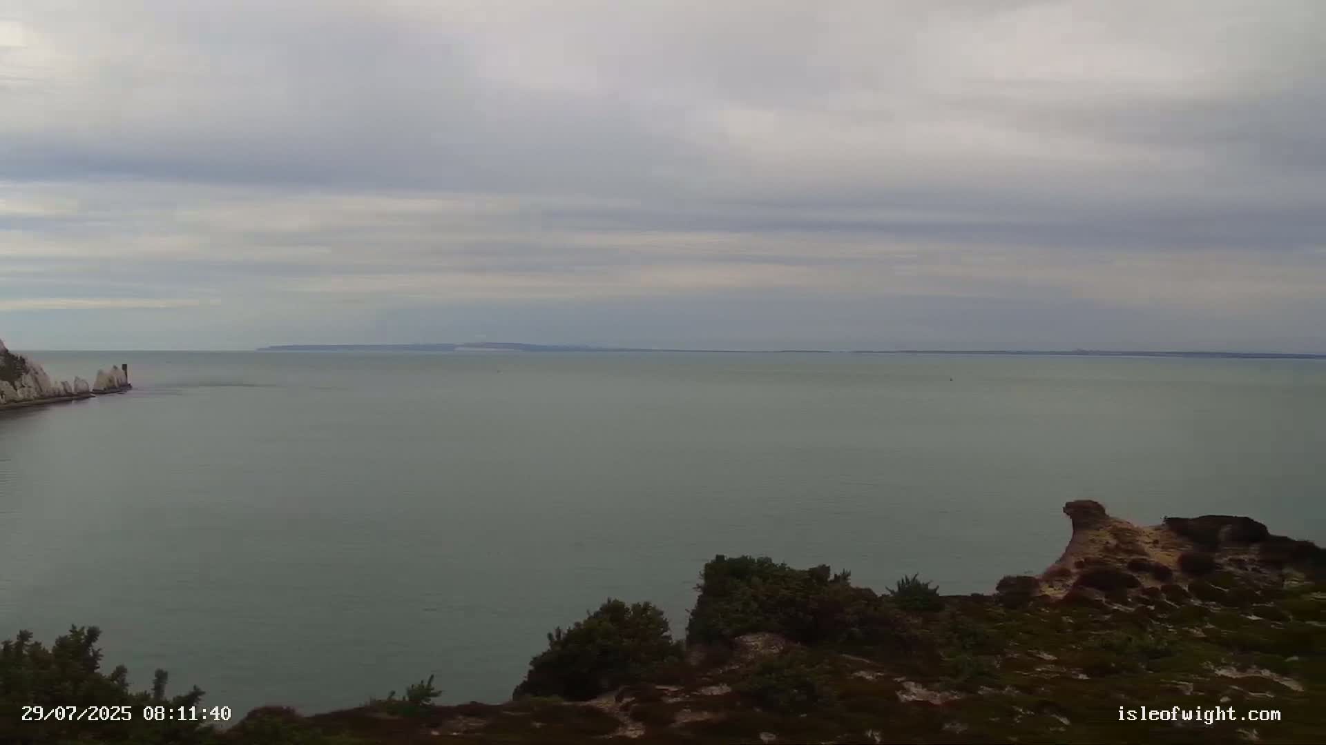 A calm, overcast day reveals a wide expanse of sea, with rocky, shrub-covered headlands in the foreground and a distant coastline visible on the horizon.