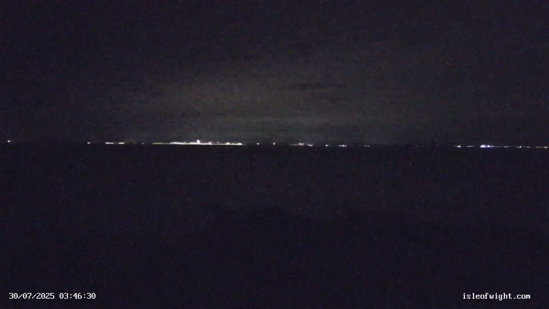 A calm, overcast day reveals a wide expanse of sea, with rocky, shrub-covered headlands in the foreground and a distant coastline visible on the horizon.
