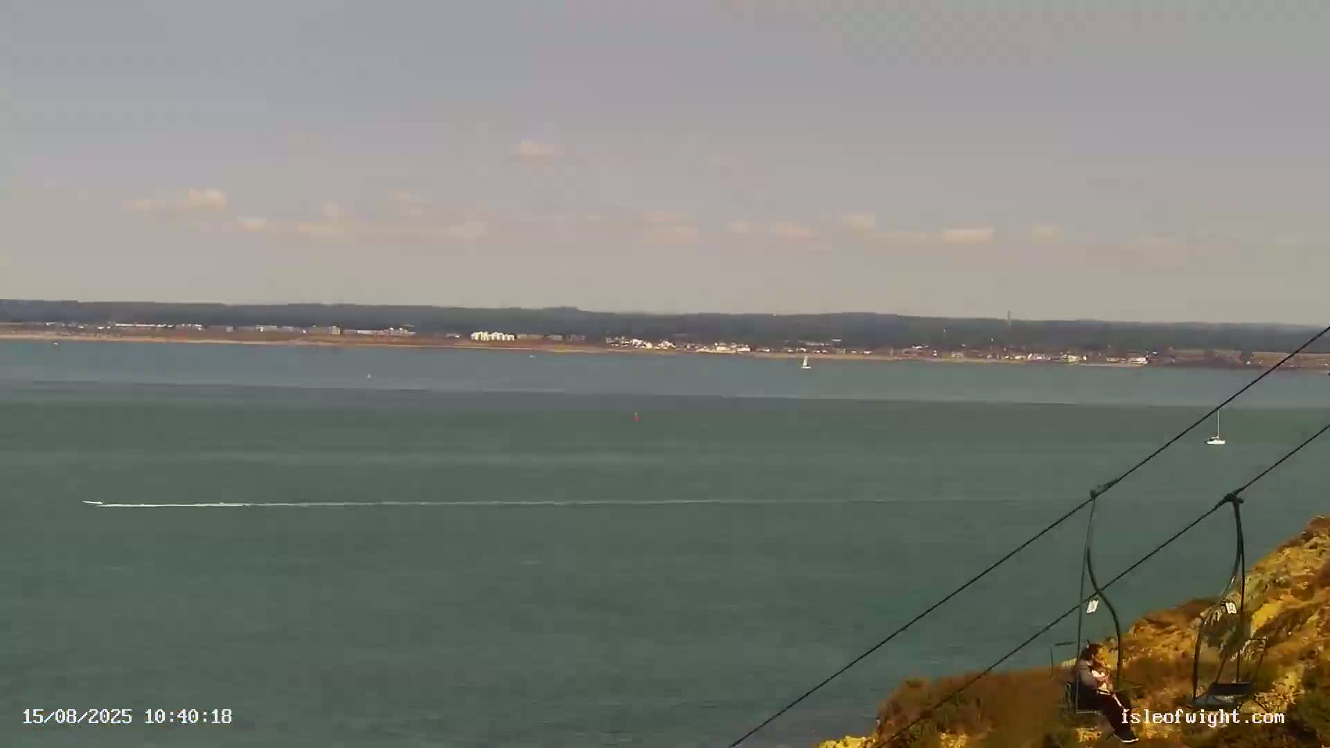 A calm, overcast day reveals a wide expanse of sea, with rocky, shrub-covered headlands in the foreground and a distant coastline visible on the horizon.