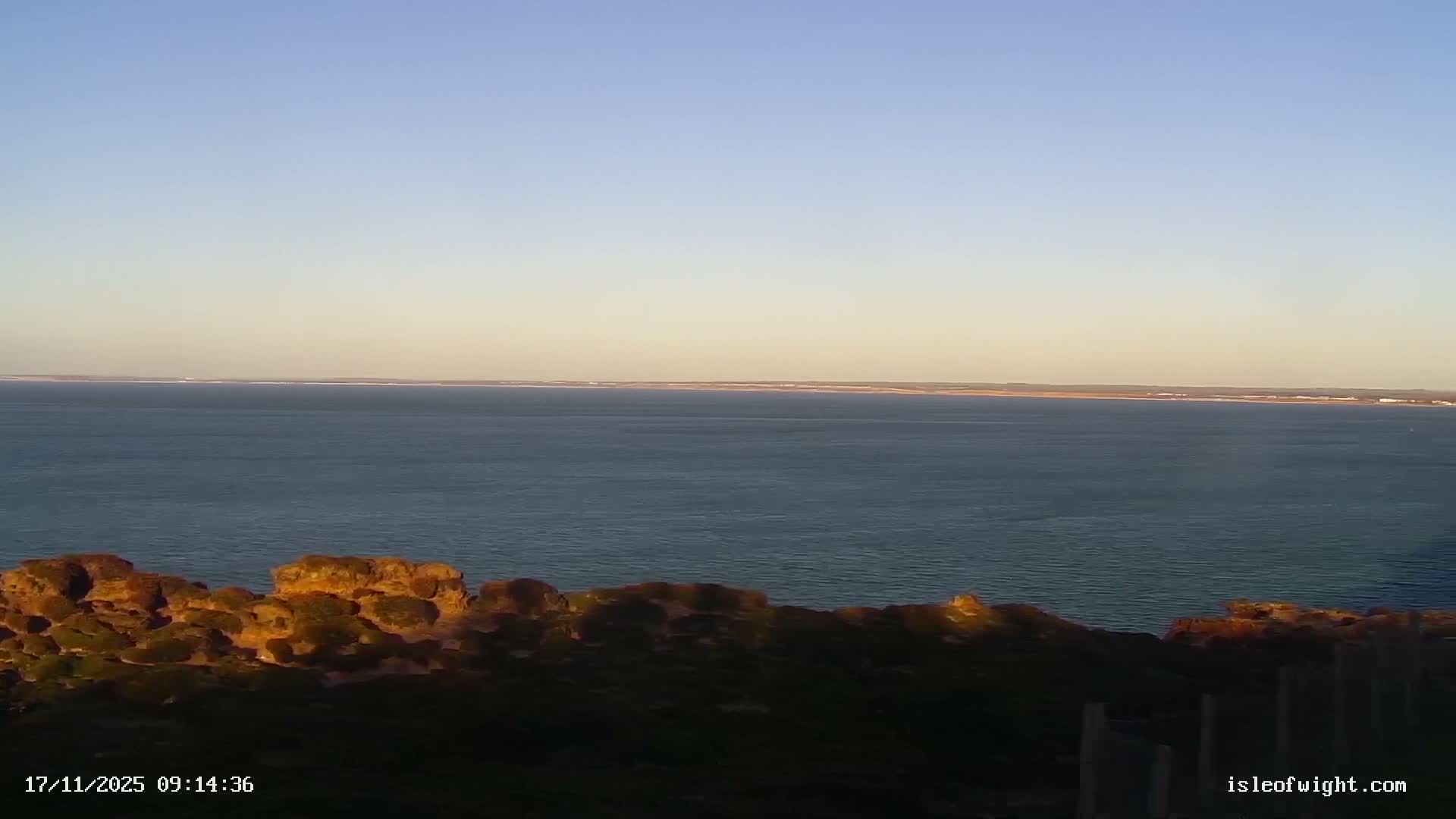 A tranquil, sunny morning view across a calm expanse of sea, framed by sunlit rocky cliffs in the foreground and a distant land strip, all under a clear sky fading from pale blue to a warm yellow glow at the horizon.