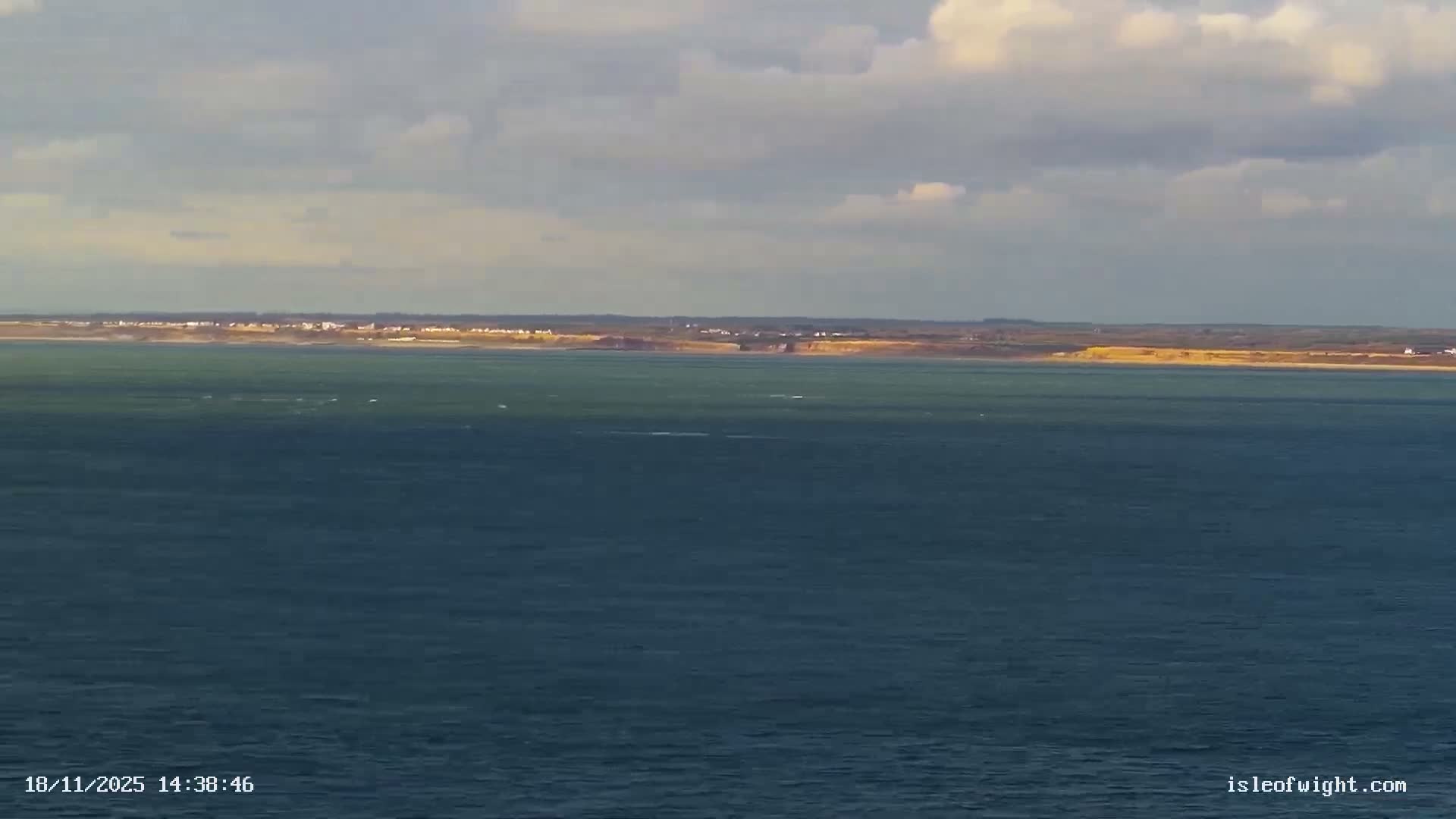 A tranquil, sunny morning view across a calm expanse of sea, framed by sunlit rocky cliffs in the foreground and a distant land strip, all under a clear sky fading from pale blue to a warm yellow glow at the horizon.
