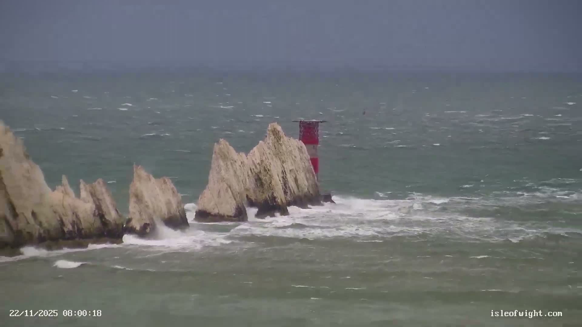 A red and white striped lighthouse stands amidst a series of rugged chalk stacks in rough, dark green-blue ocean waters with crashing waves under an overcast, stormy sky.