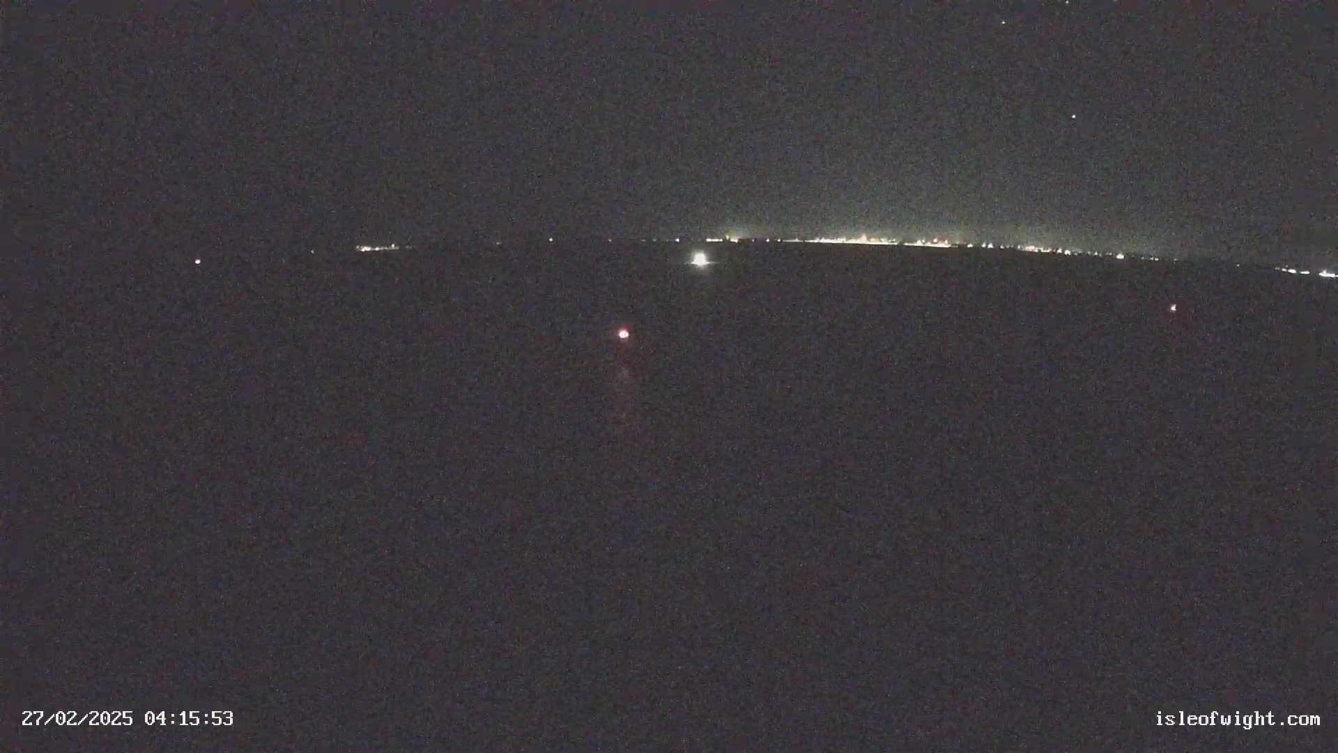 A nighttime, dark scene shows a distant line of lights on the horizon and a few small lights closer in the foreground under a clear, starry sky.