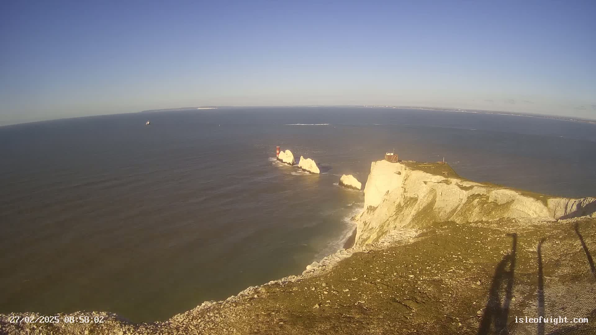 A high-angle view shows a rocky coastline and a cluster of white rock formations in the sea under a clear blue sky.