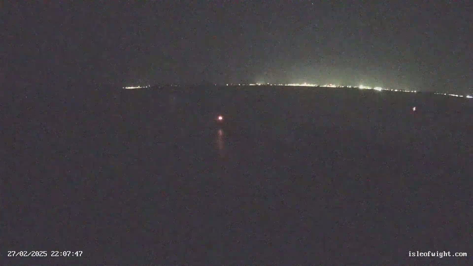 A dark, nighttime scene shows a distant line of lights on the horizon and a single, small, reddish light closer to the camera in the water.