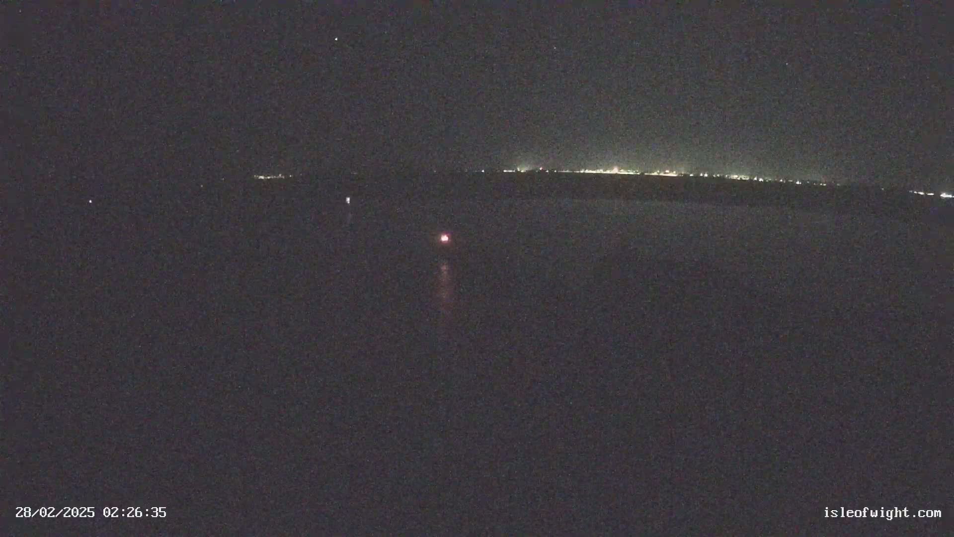 A dark, nighttime scene shows a distant line of lights on the horizon above a dark body of water with a single, small, faint reddish light visible closer to the camera.