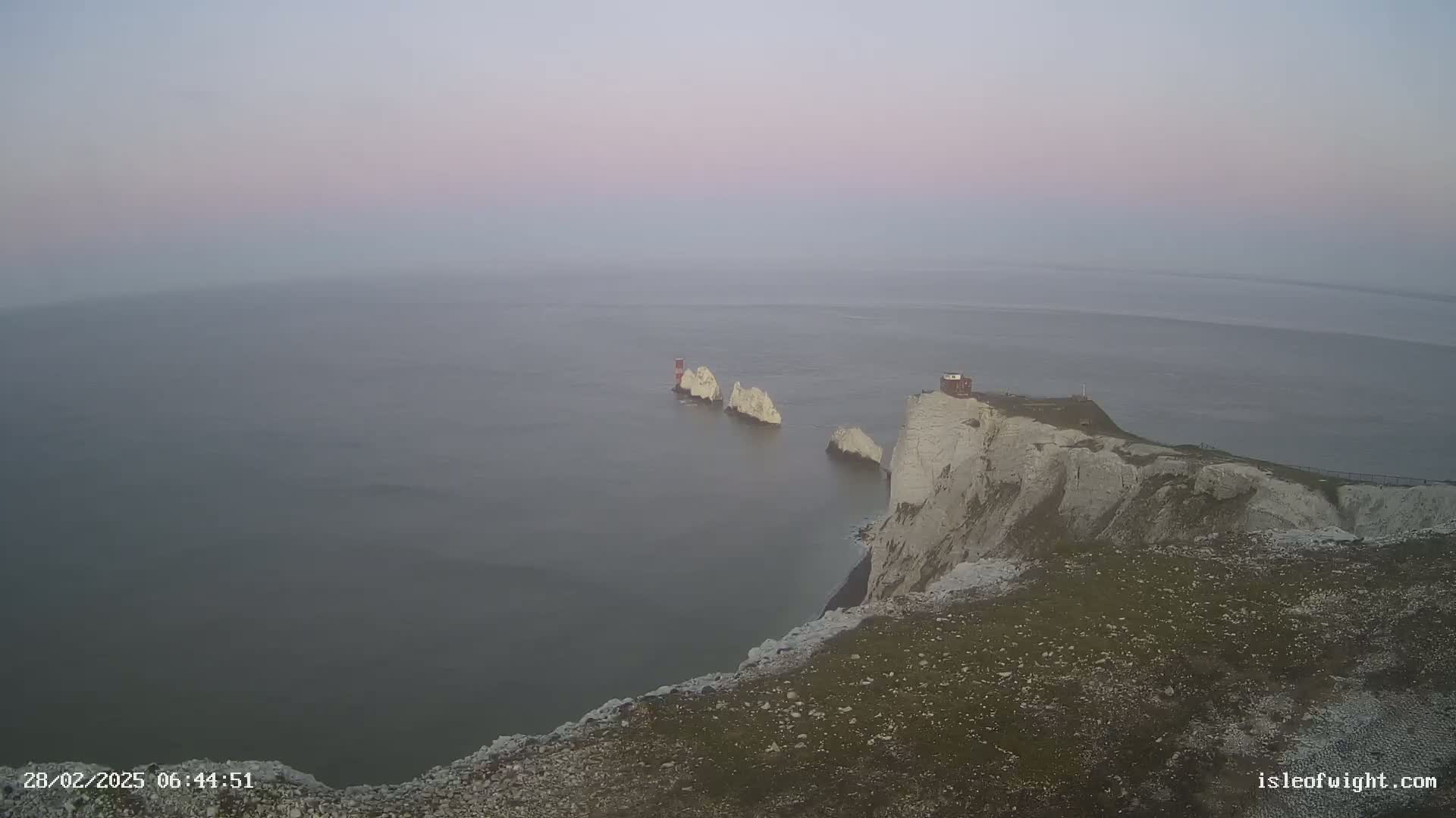 A hazy, pale sunrise illuminates white cliffs overlooking a calm sea and several smaller, distant white rock formations.