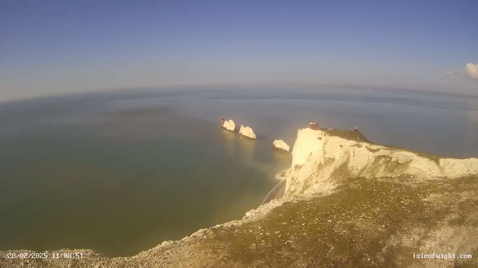 A high-angle view shows a cliff overlooking a calm sea with several stacks of white rock formations in the distance under a clear blue sky.