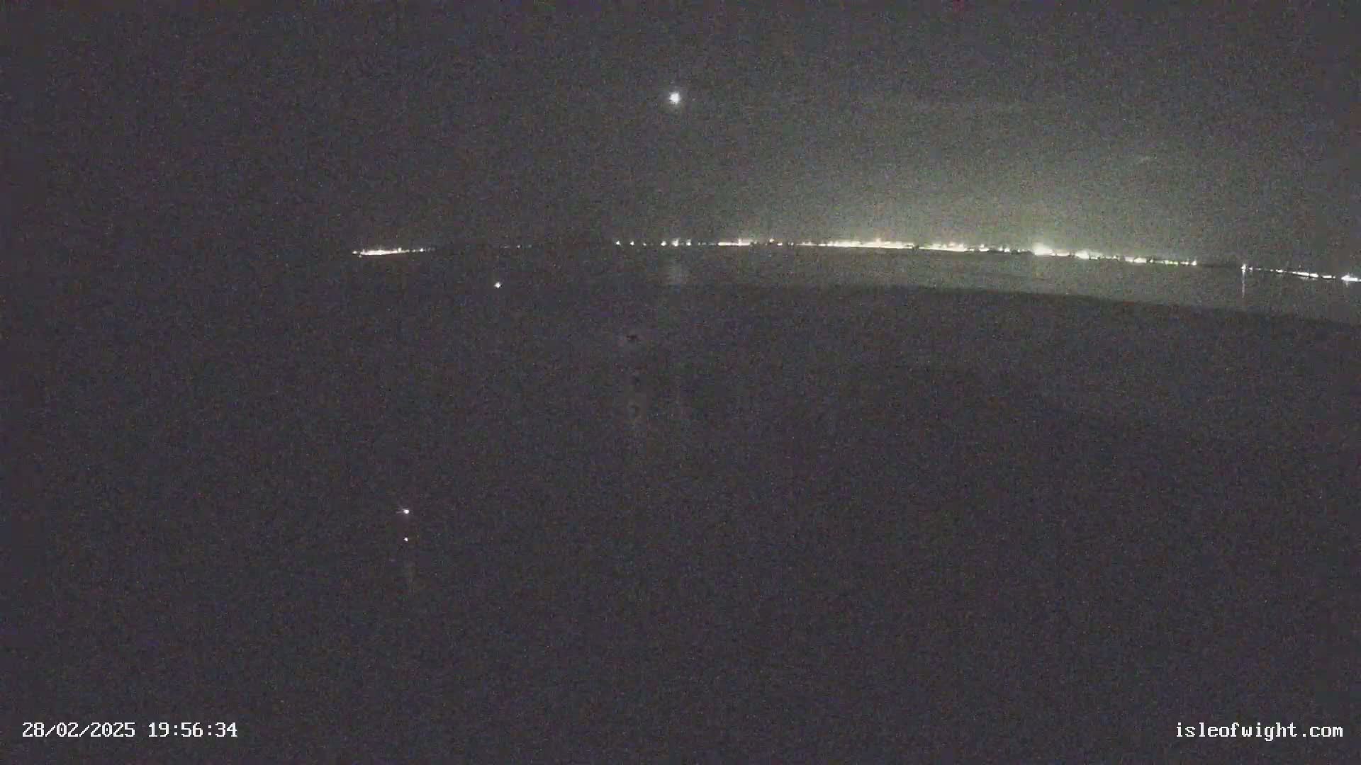 A nighttime, long-range view of a dark body of water reflecting a distant line of lights under a clear, dark sky.