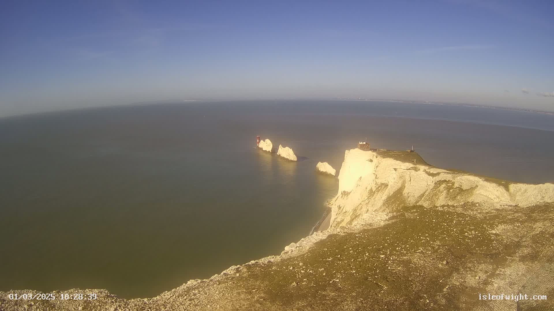 High-angle view of a coastal landscape on a clear, sunny day, showing white cliffs meeting a calm sea with several rock formations extending into the water.