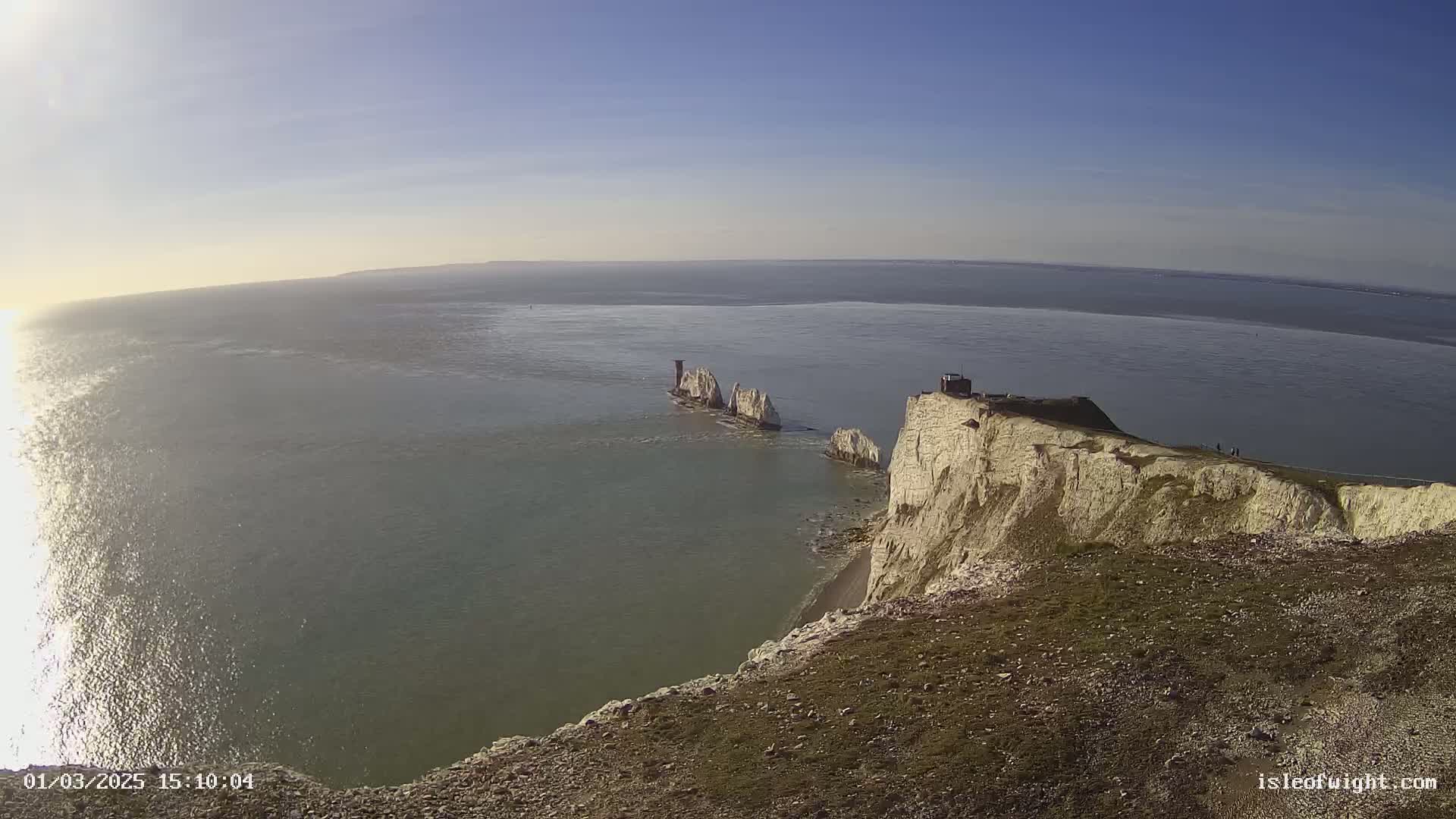 A bright, sunny day reveals a panoramic view of white cliffs overlooking a calm sea, with a distant group of smaller rock formations visible in the water.