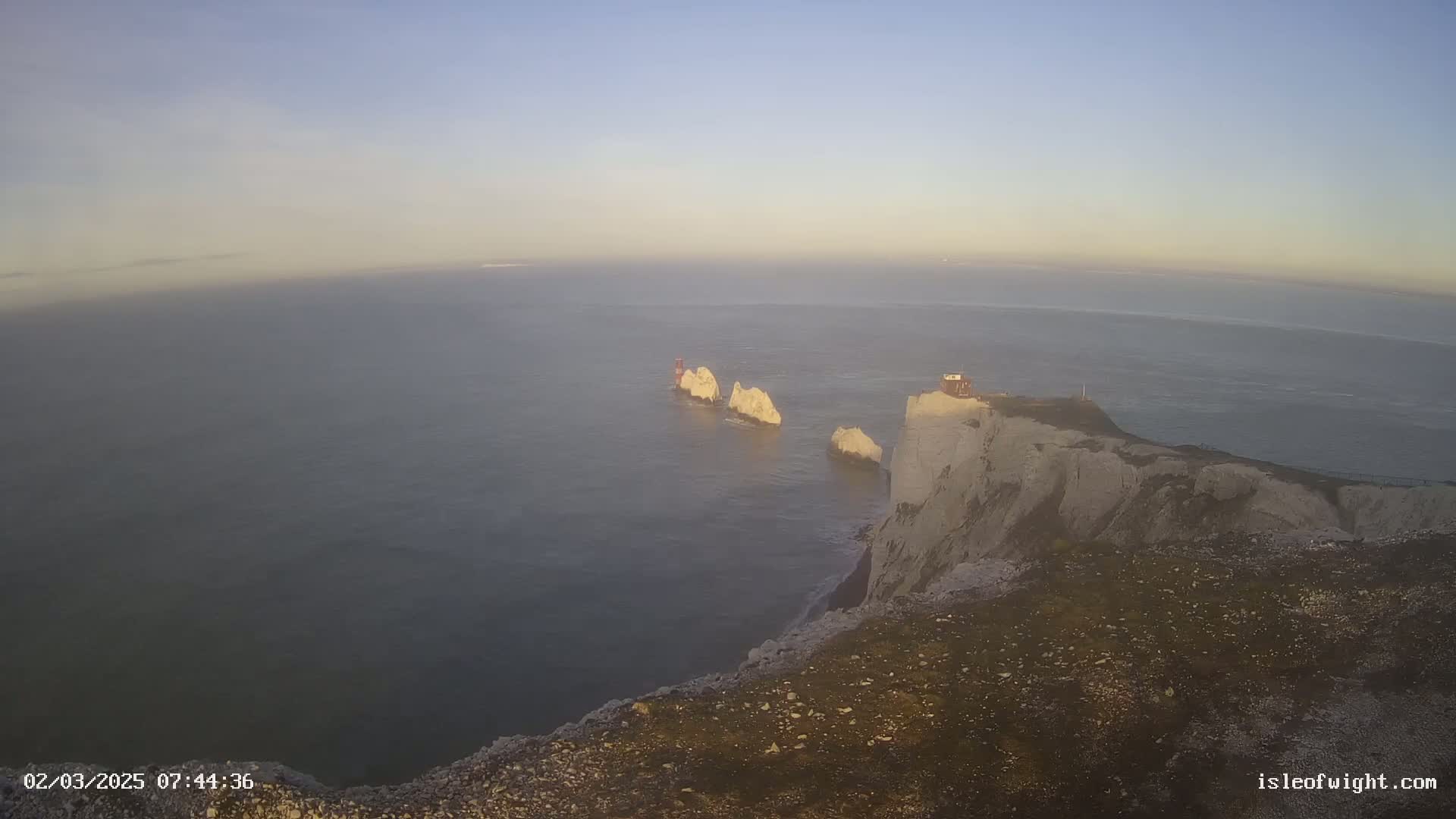 A high-angle view shows a calm sea under a clear, light-blue sky, with white cliffs and several stacks of rock formations visible near the shore.