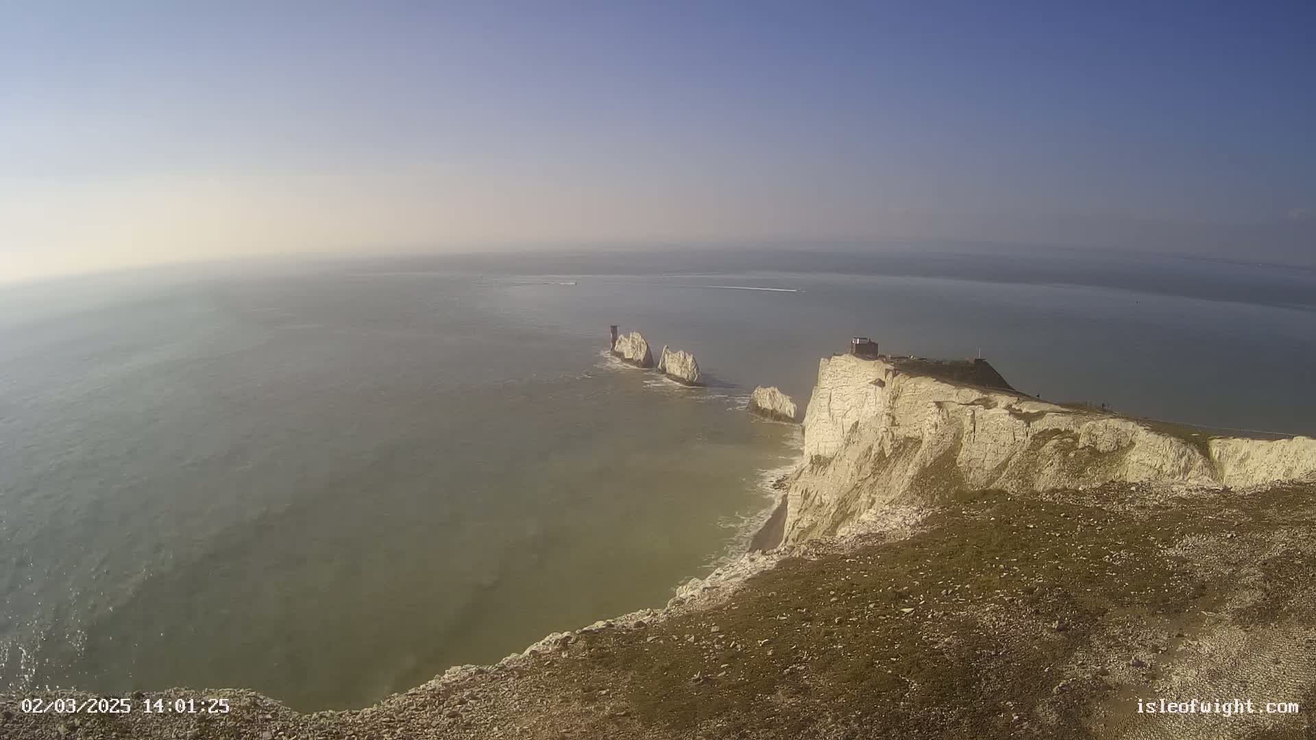 A high-angle view shows a rocky, white cliff overlooking a calm, hazy sea where several stacks of rock jut out from the water.