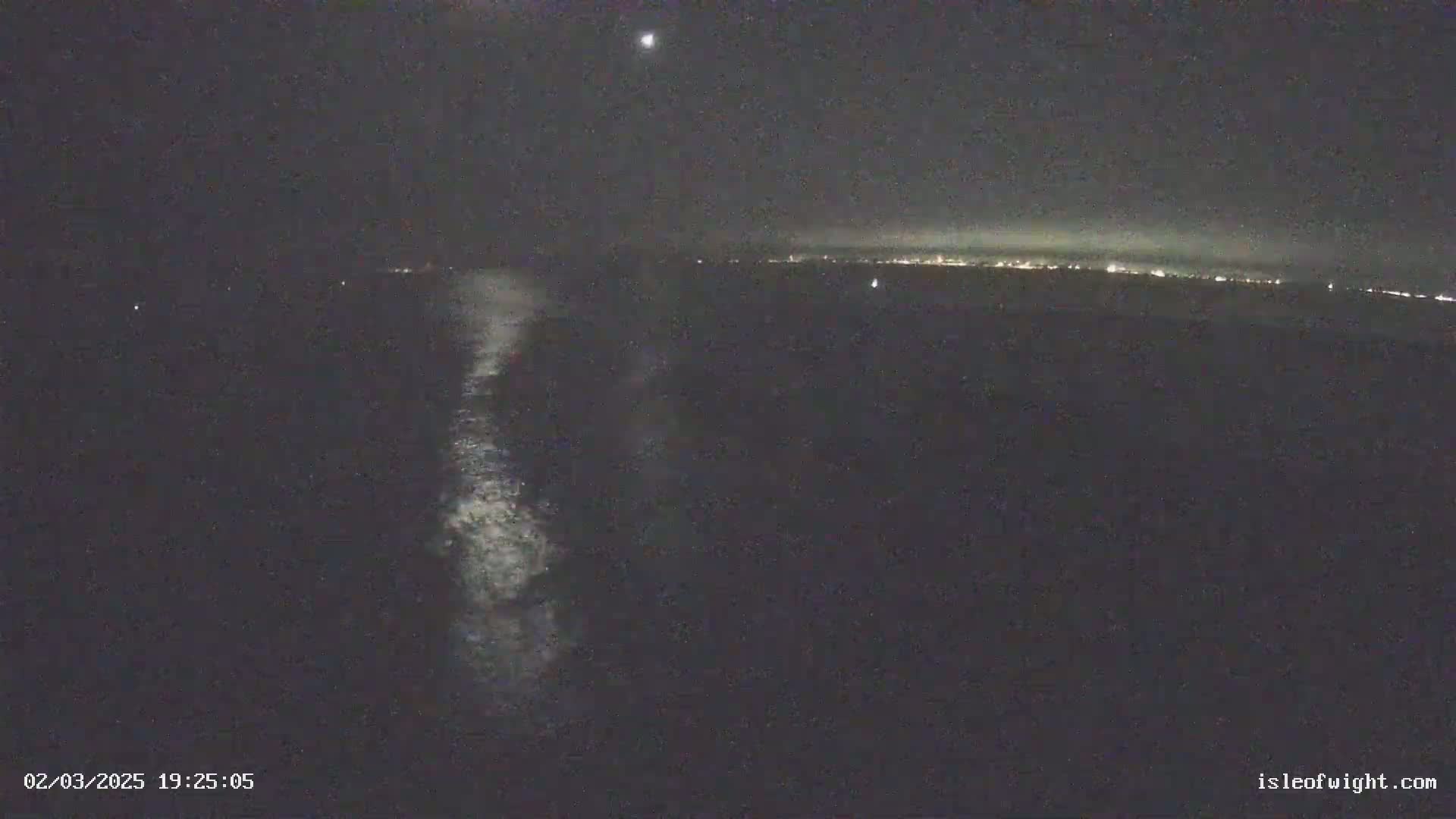 A nighttime view of a dark body of water with a distant, faintly lit shoreline and a bright wake trailing behind a vessel.