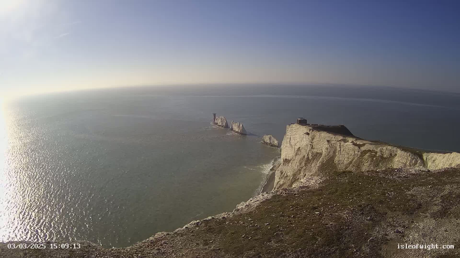 A high-angle view shows white cliffs meeting a calm, sunlit sea, with several stacks of rock visible in the water.
