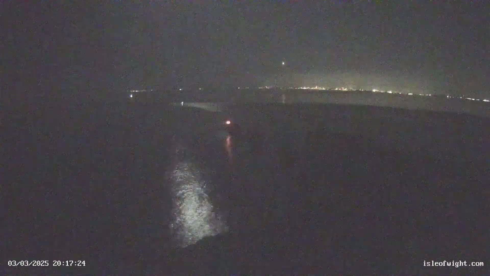 A nighttime, low-light view shows a dark body of water with faint wave action, a small light in the distance, and a distant shoreline with faint city lights.