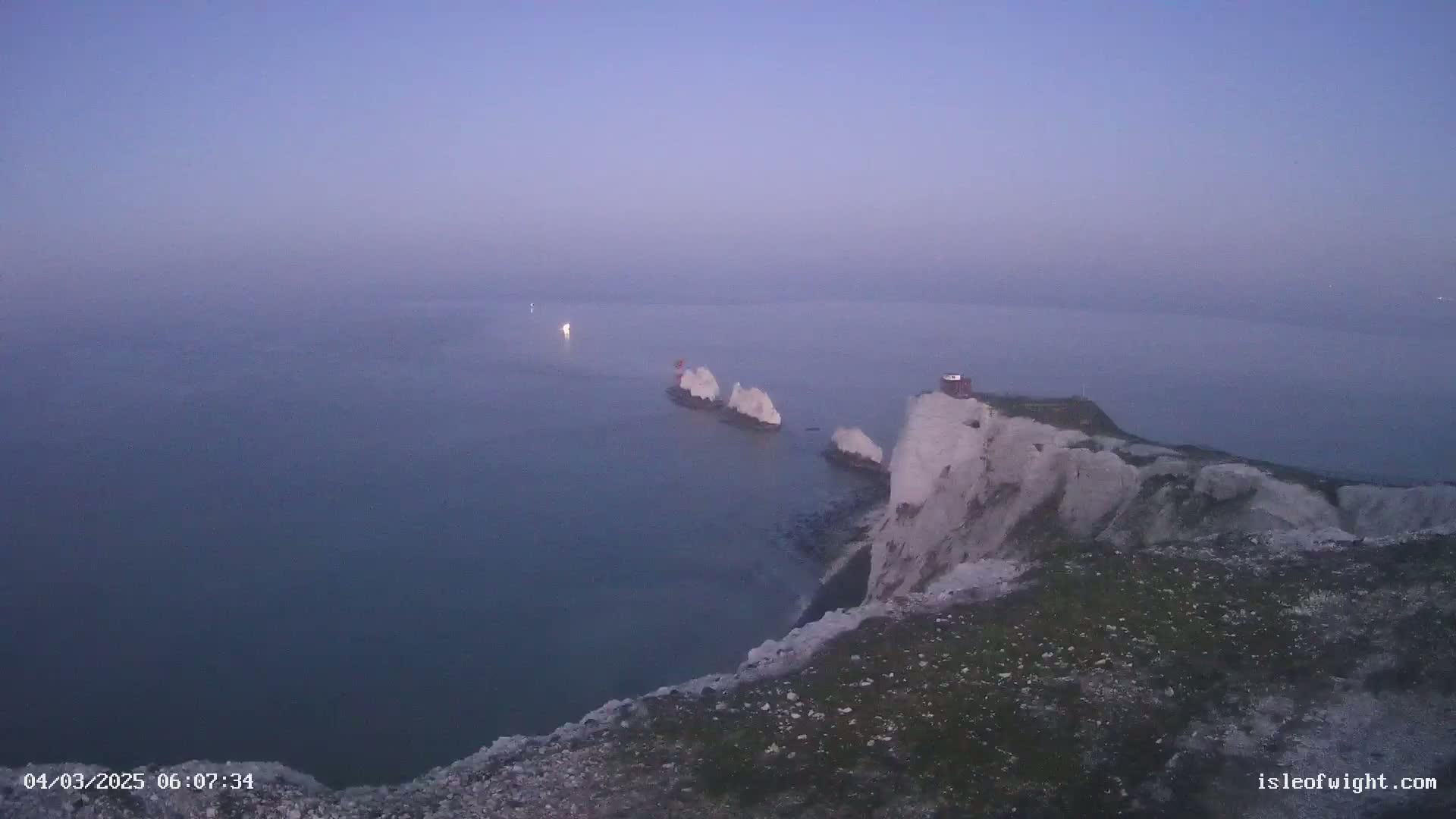 A hazy, pre-dawn view shows white cliffs overlooking a calm sea where several small, white rock formations are visible.