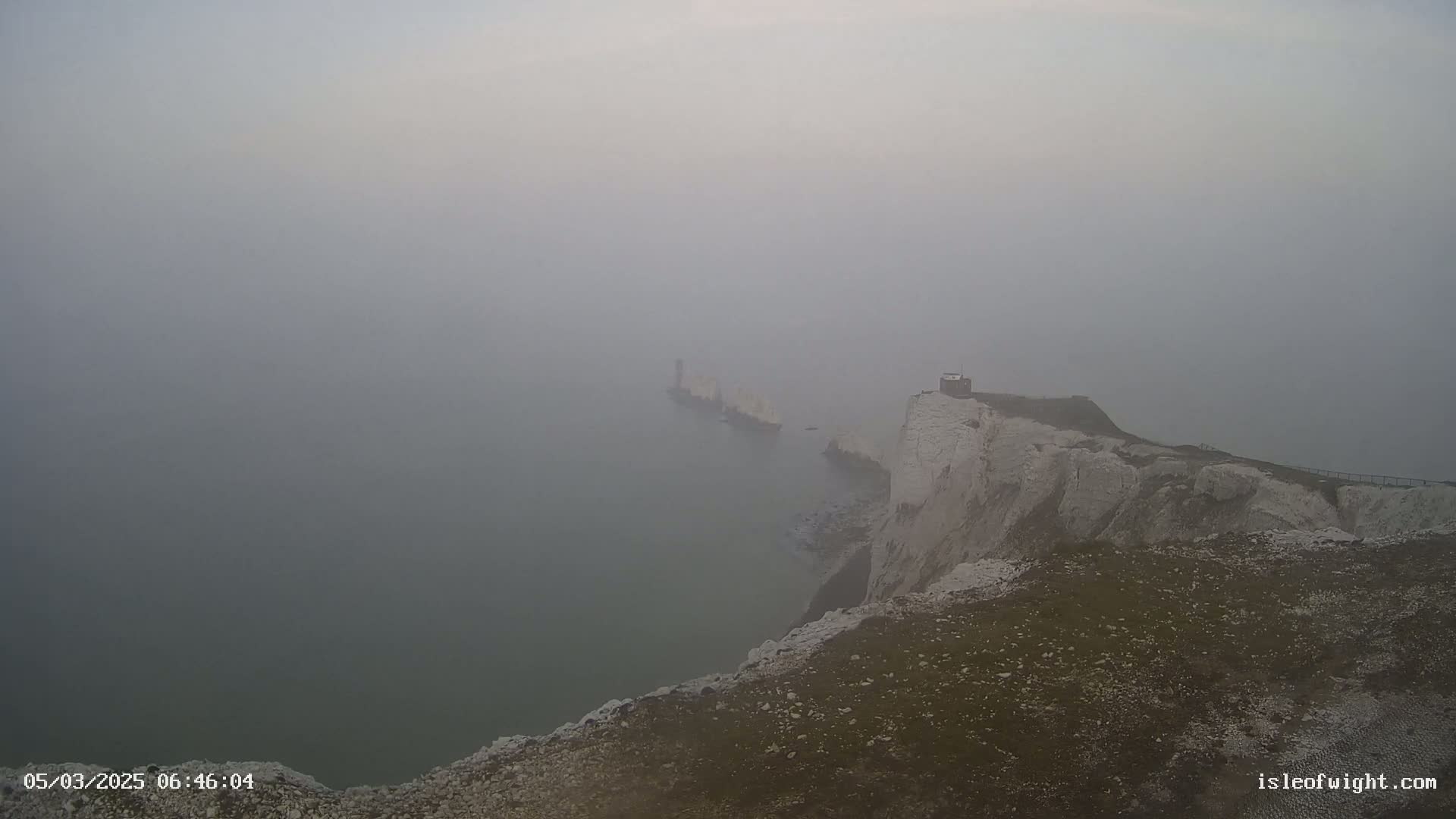 A foggy view shows white cliffs descending to a hazy sea, with a structure visible on the cliffs and another in the distance across the water.