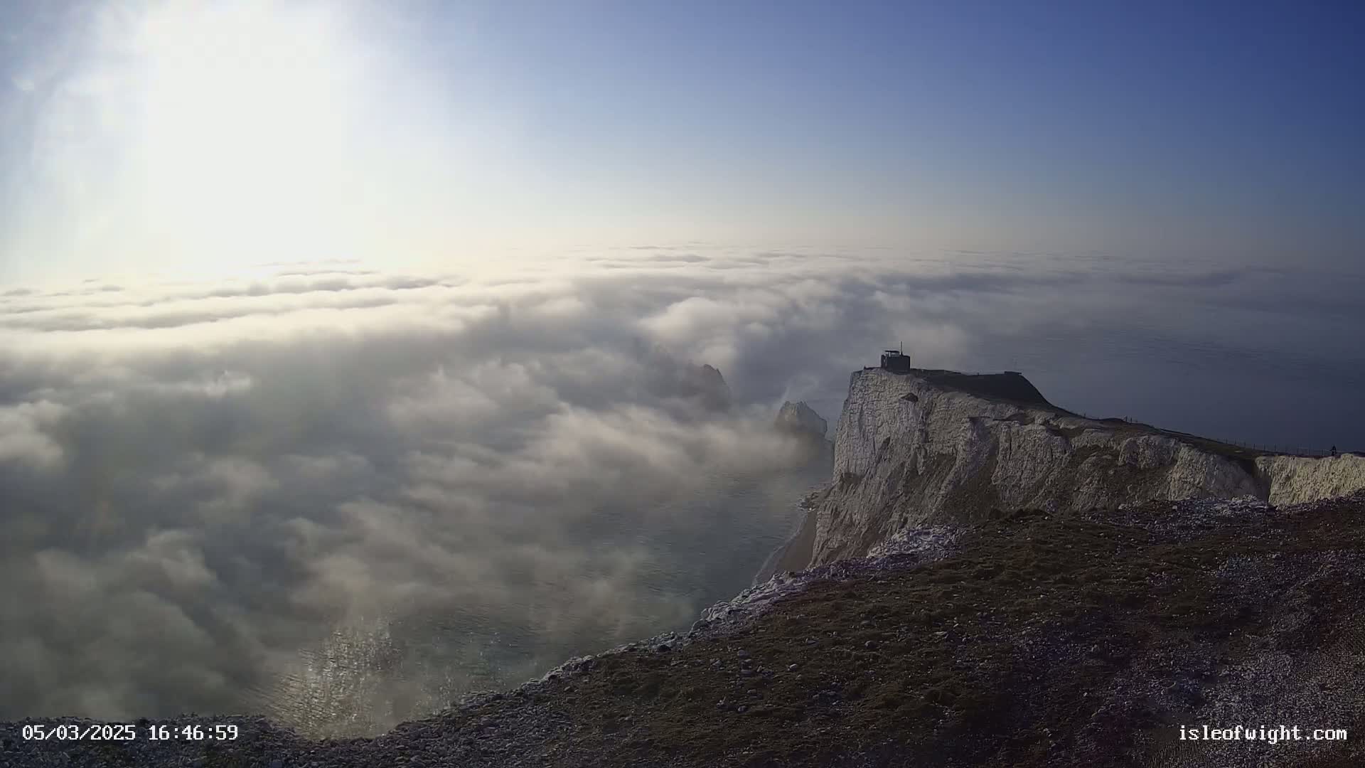 A high vantage point overlooking a dramatic white cliff face and the sea, shrouded in a thick layer of low-lying clouds under a mostly clear sky.