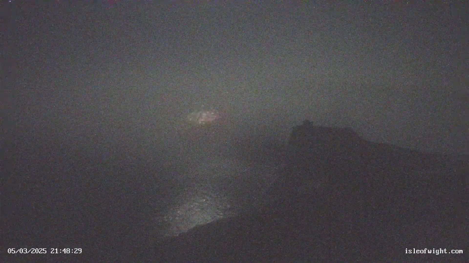 A hazy, dark night scene shows a faint, light-colored object in the distance above a dark, silhouetted hill.