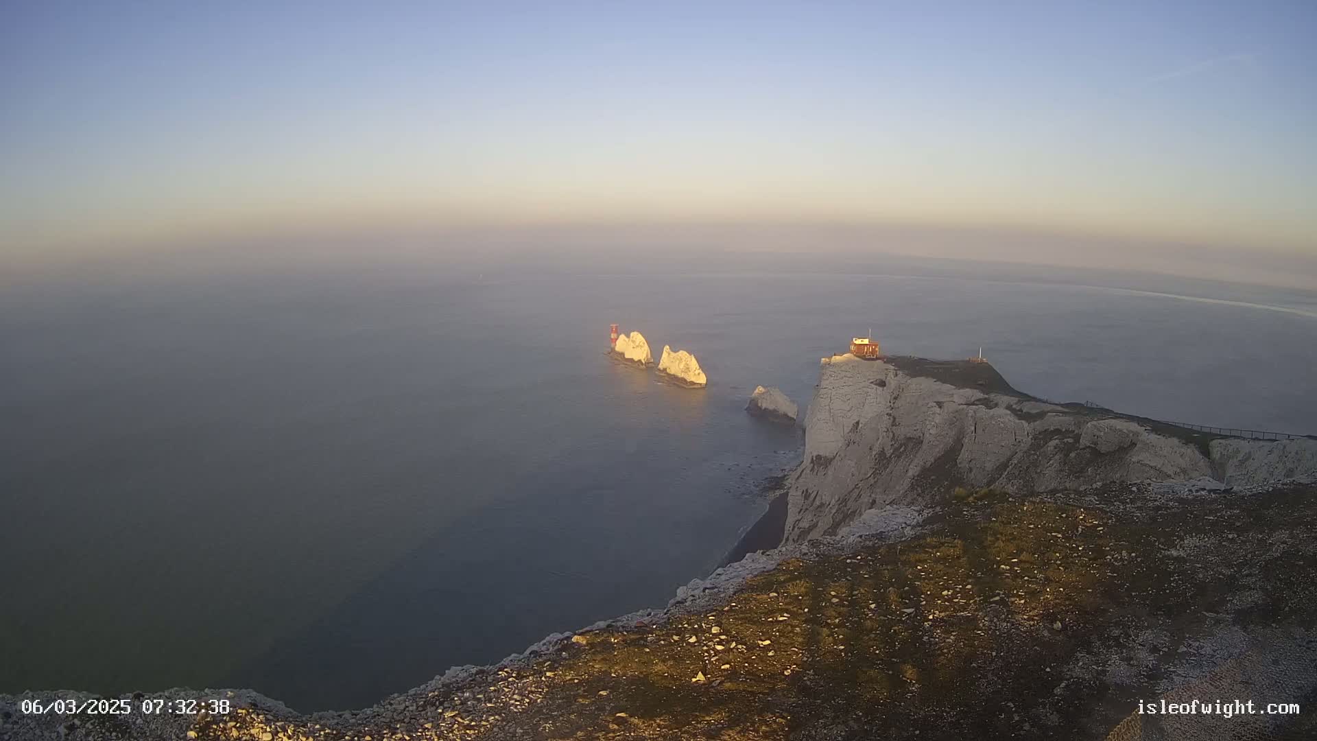 White cliffs overlook a calm, foggy sea where several smaller white rock formations and a small red structure are visible in the distance.