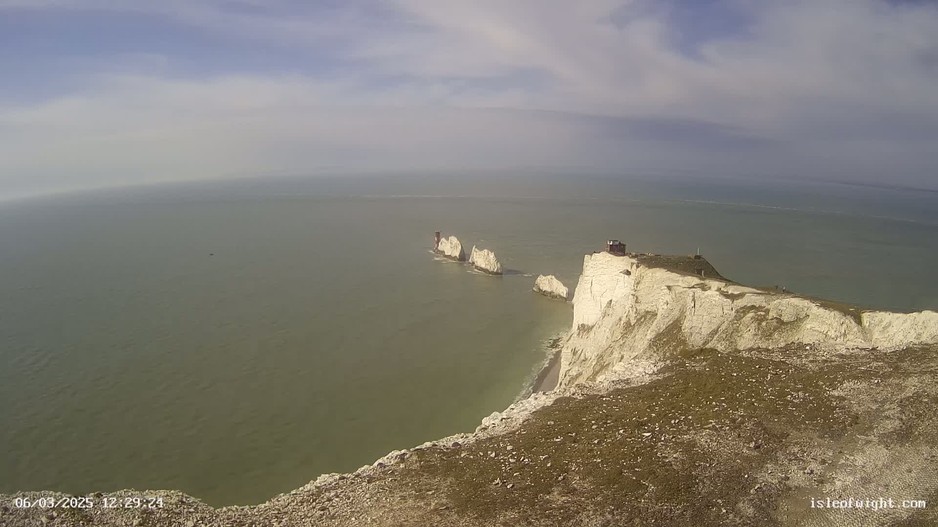 White cliffs overlooking a calm sea with a chain of smaller white rock formations extending into the water under a partly cloudy sky.