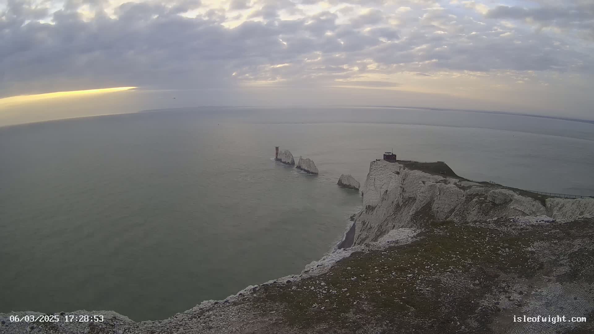 A high-angle view of a cliff overlooking a calm sea with several white rock formations in the distance under a cloudy sky.