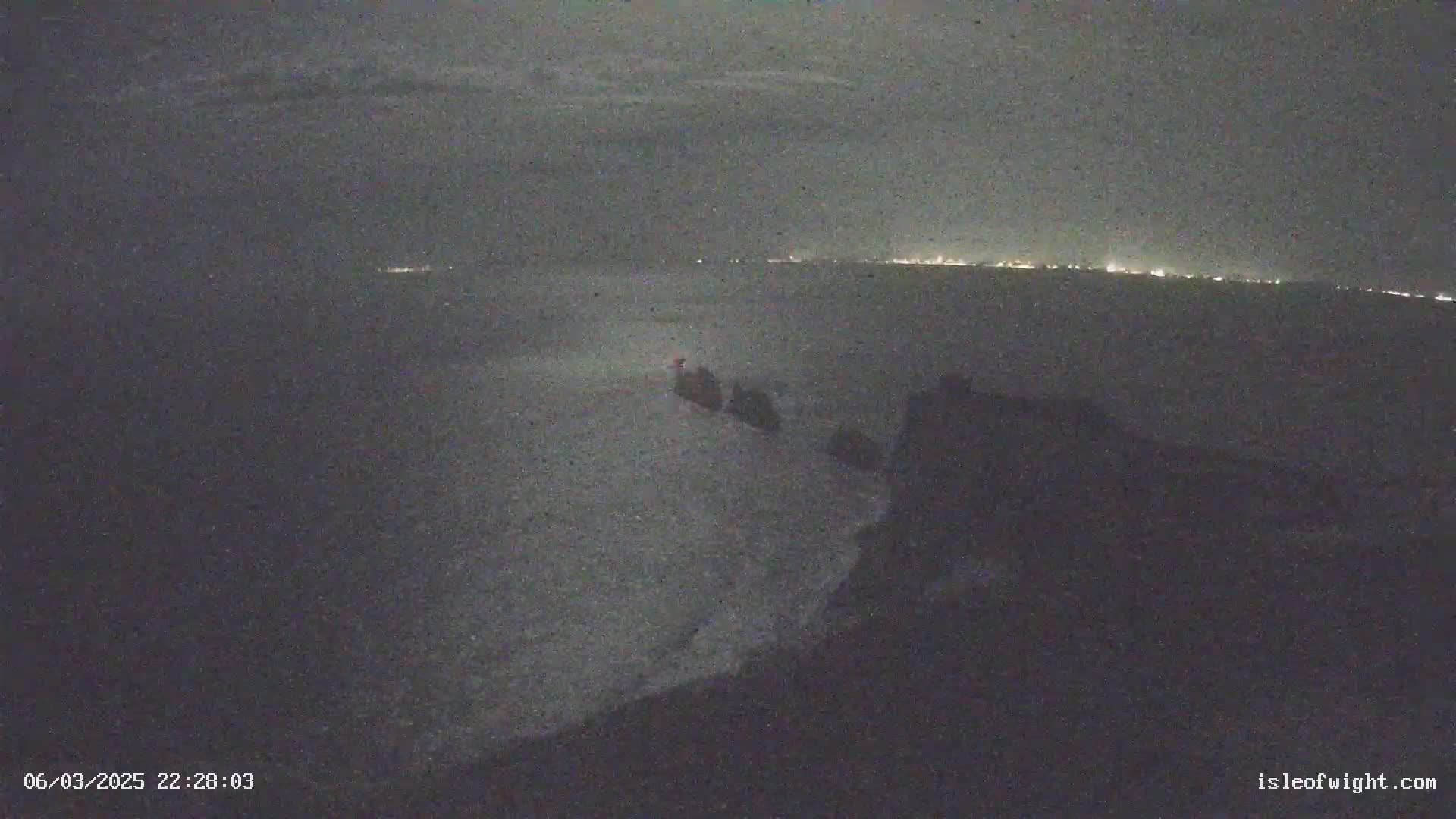 A nighttime coastal scene shows dark, wavy water with a distant line of lights on the horizon under a cloudy sky.