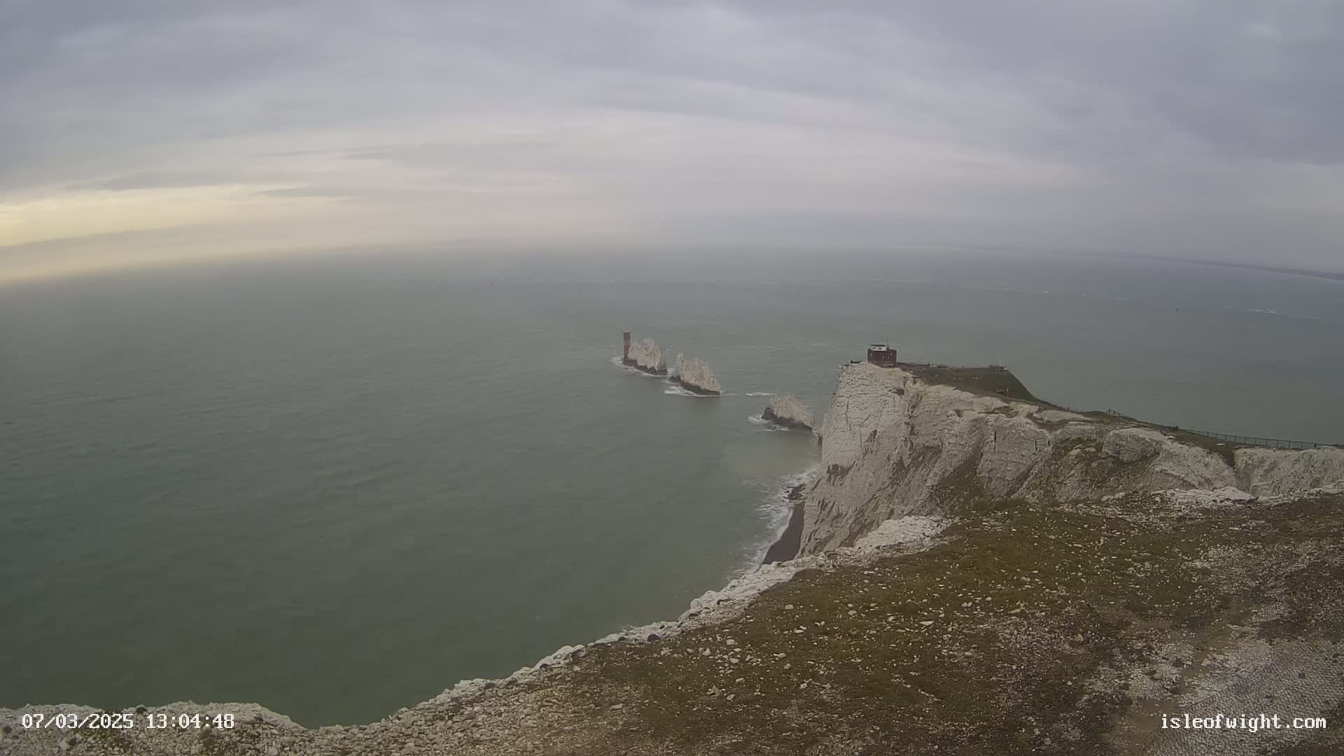 White cliffs overlooking a gray, calm sea with several smaller white rock formations visible in the distance under an overcast sky.
