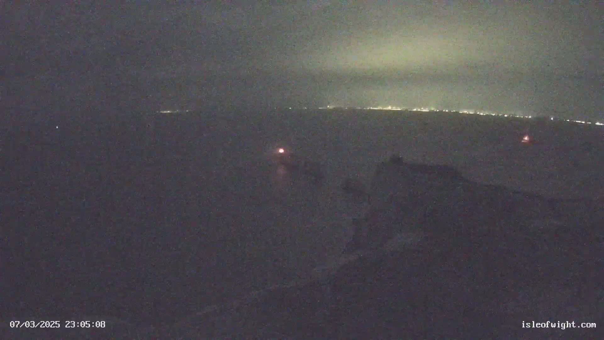A nighttime, low-light view of a body of water shows a distant shoreline with lights, and a few faint lights on the water closer to the camera.