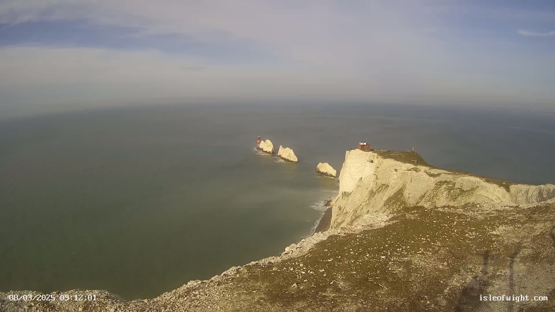 A high-angle view shows white cliffs meeting a calm, gray-green sea, with a line of smaller, distant white rock formations.