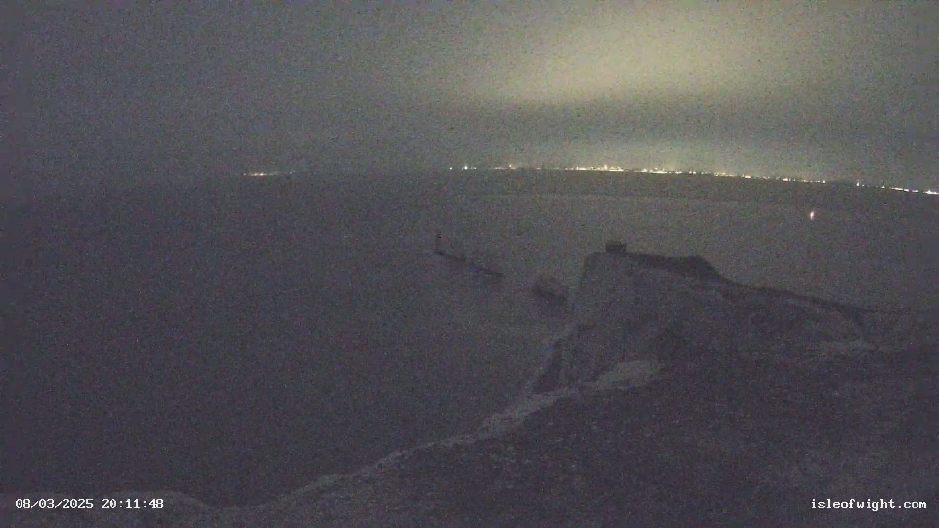 A nighttime, overcast view from a high vantage point shows a dark body of water with a distant city skyline faintly illuminated, and a rocky, dark foreground.