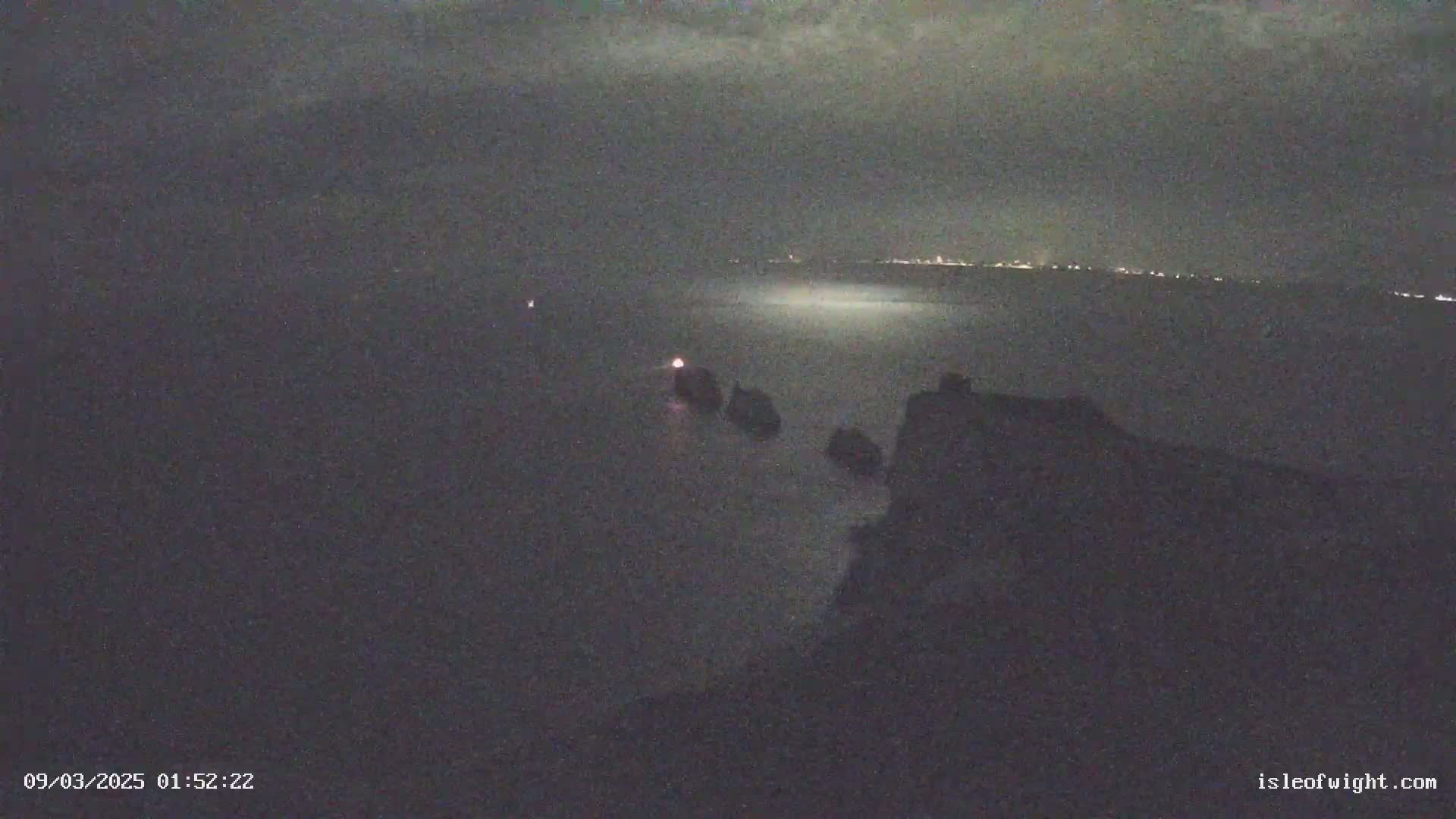 A nighttime, low-light view of a dark, rocky coastline with a body of water extending to a faintly lit horizon.