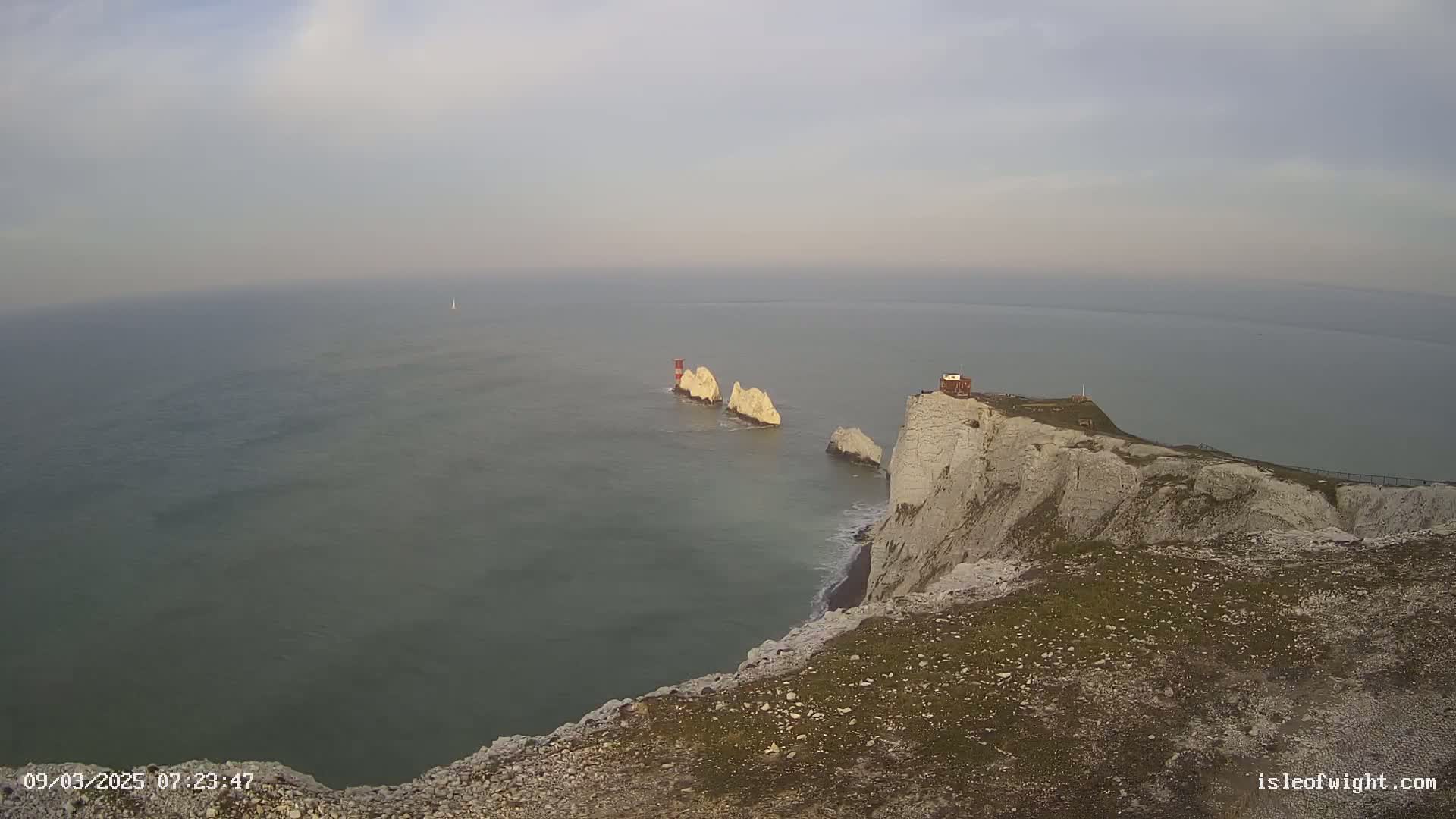 A high-angle view shows white cliffs overlooking a calm sea with several small white rock formations and a small building on the clifftop, under an overcast sky.