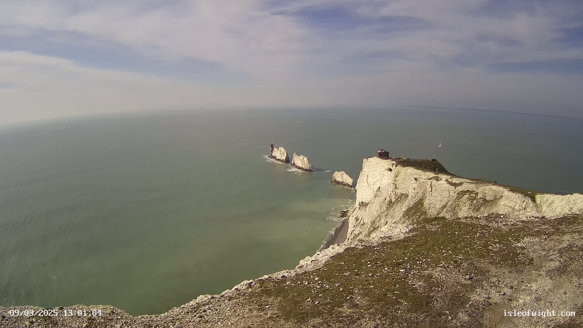White cliffs overlooking a calm, green-gray sea with a line of smaller white rock formations and a lighthouse in the distance under a partly cloudy sky.