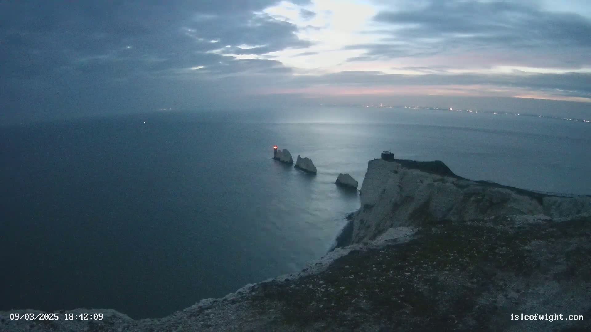 A dark, overcast sky over a body of water with white cliffs and stacks, one of which has a light on top.