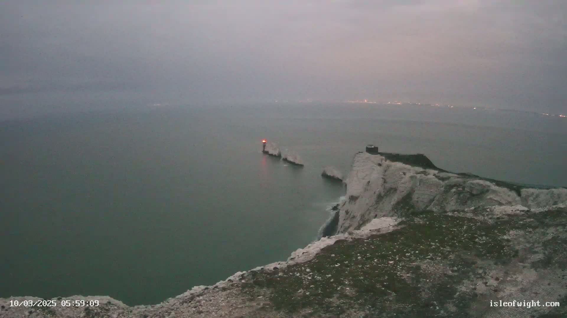 White cliffs overlook a calm, grey sea with a distant lighthouse illuminated by a red light.
