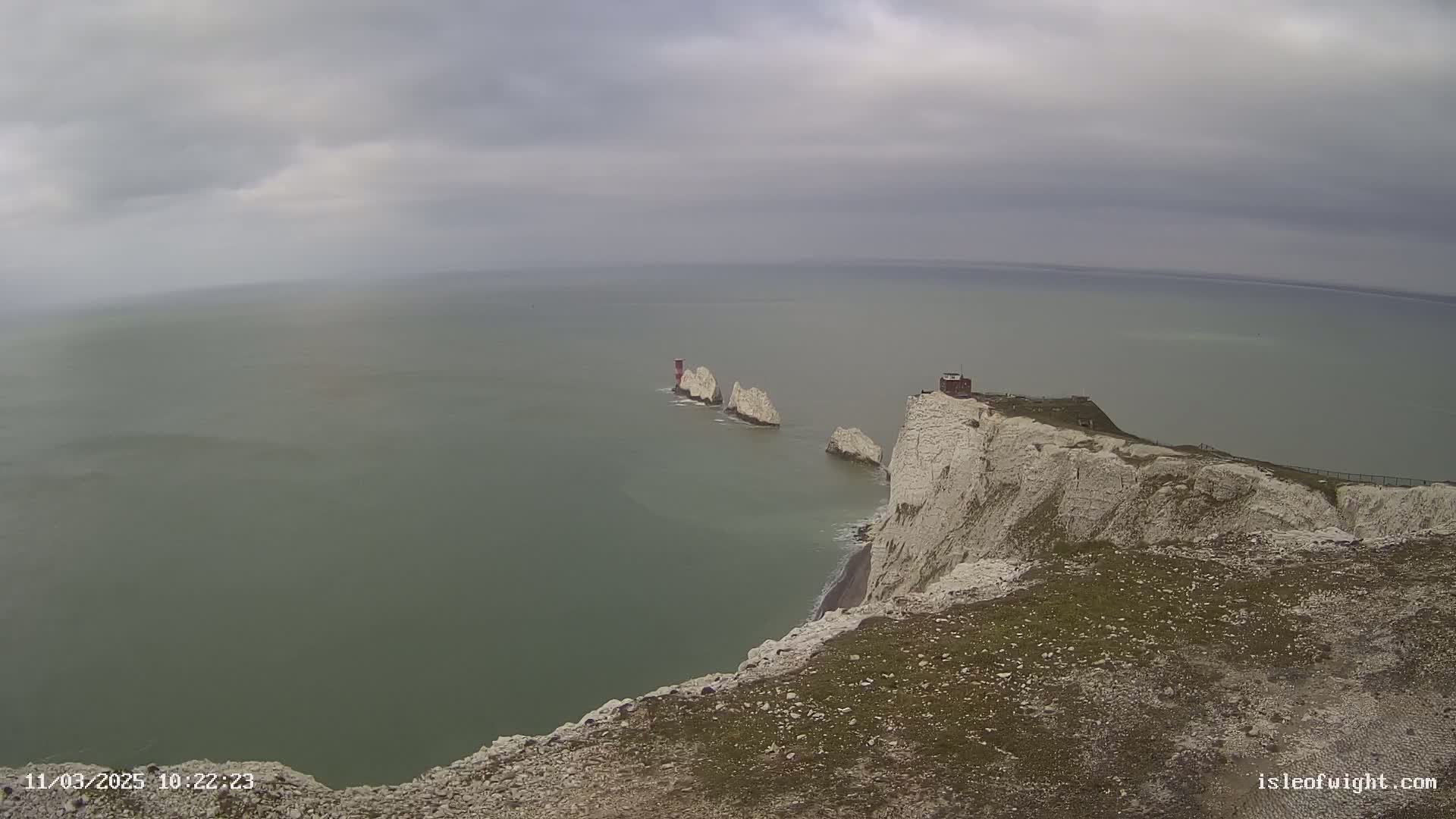 White cliffs overlooking a calm, gray-green sea with several smaller white rock formations and a small building visible in the distance under an overcast sky.