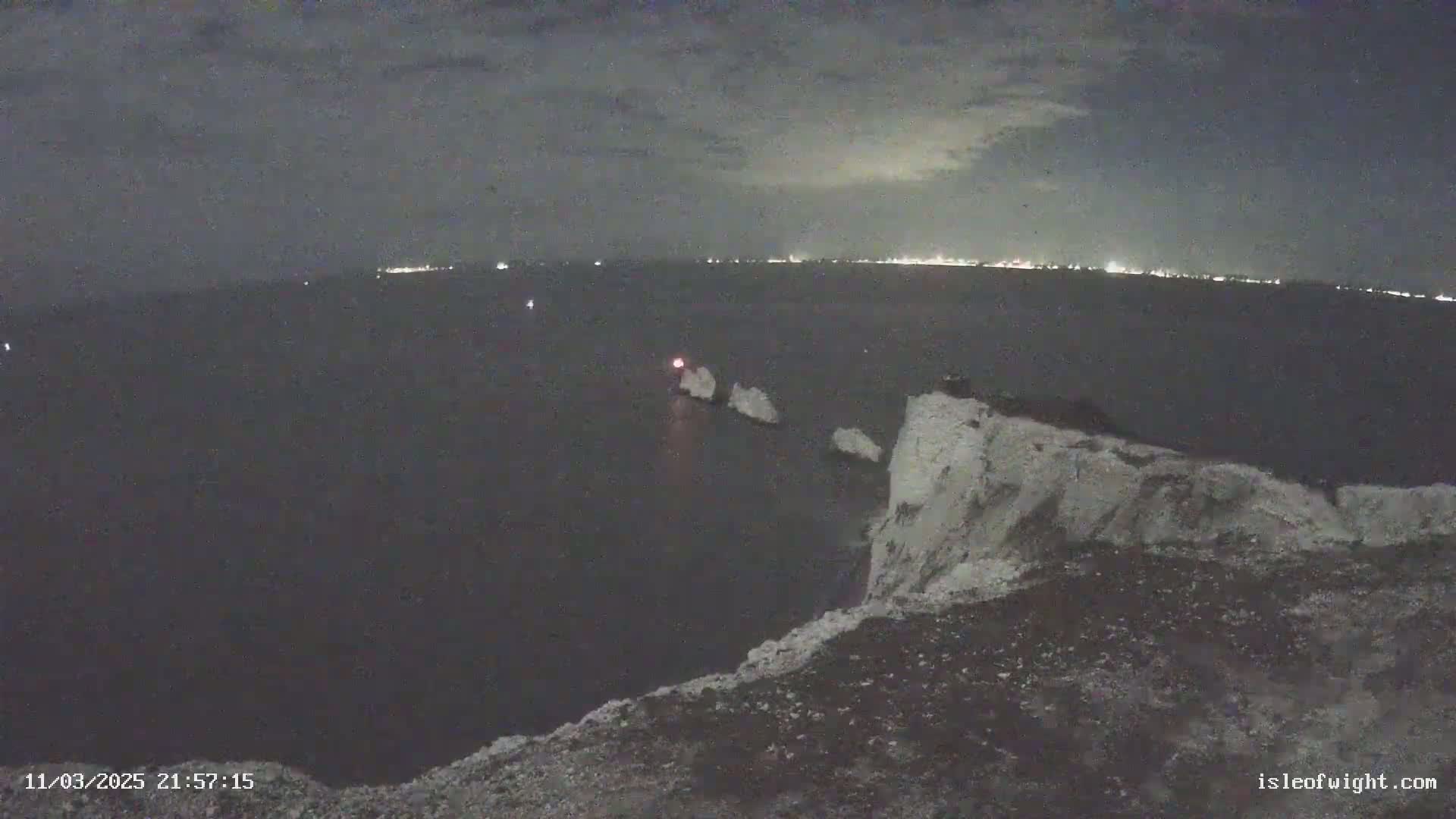 A nighttime view from a cliff overlooking a dark sea with distant city lights and a few small, illuminated rocks in the water.