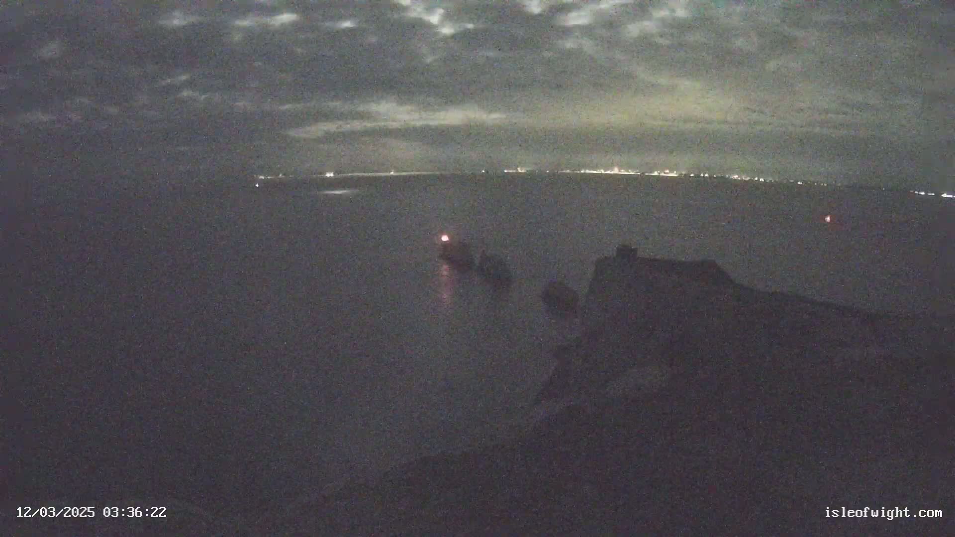A nighttime, cloudy view of a dark body of water with a few lights visible on the horizon and a small light on a vessel in the foreground.